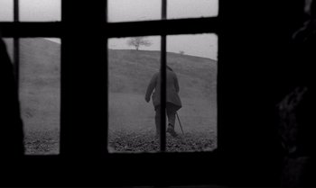 Movie still from “The Turin Horse” (2011), directed by Ágnes Hranitzky – A man standing on a dirt field looking out a window; Wide shot, Low angle