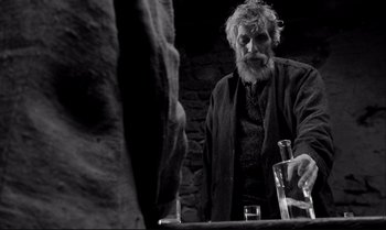 Movie still from “The Turin Horse” (2011), directed by Ágnes Hranitzky – An old man with a long white beard is pouring water into a glass; Medium shot, Over the shoulder angle