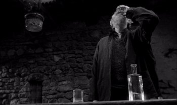Movie still from “The Turin Horse” (2011), directed by Ágnes Hranitzky – An old man standing in front of a table with two glasses; Medium shot, Low angle