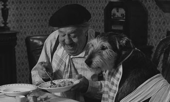 Movie still from “The Two of Us” (1967), directed by Claude Berri – An old man and a dog are sharing a meal together; Medium shot, High angle