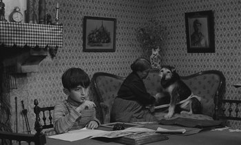 Movie still from “The Two of Us” (1967), directed by Claude Berri – A boy sitting at a table with papers and a dog; Medium shot, High angle