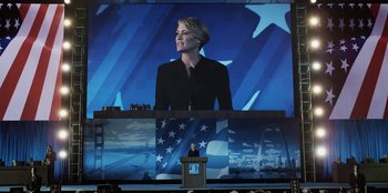 Movie still from “House of Cards” (2013), created by Beau Willimon – A woman standing in front of an american flag on a large screen; Wide shot, Low angle