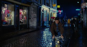 Movie still from “The Umbrellas of Cherbourg” (1964), directed by Jacques Demy – A man riding a bike down a street at night; Wide shot, High angle