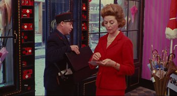 Movie still from “The Umbrellas of Cherbourg” (1964), directed by Jacques Demy – A woman in a red suit writing on a piece of paper next to a man in a uniform; Medium shot, Over the shoulder angle