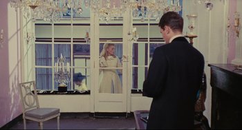 Movie still from “The Umbrellas of Cherbourg” (1964), directed by Jacques Demy – A man and a woman are standing in front of a window; Wide shot, Over the shoulder angle