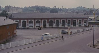 Movie still from “The Umbrellas of Cherbourg” (1964), directed by Jacques Demy – Two people riding bikes down a street next to a parking lot; Extreme Wide shot, High angle