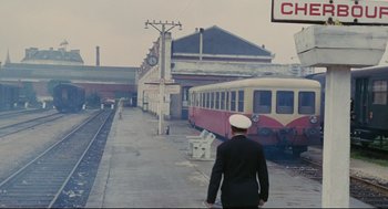 Movie still from “The Umbrellas of Cherbourg” (1964), directed by Jacques Demy – A man standing in front of a train station next to a building; Extreme Wide shot, High angle