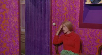 Movie still from “The Umbrellas of Cherbourg” (1964), directed by Jacques Demy – A woman sitting in a chair in front of a purple wall; Medium shot, High angle