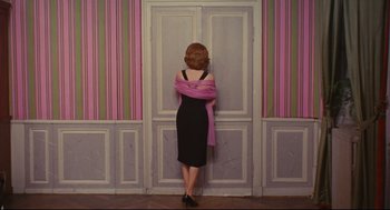 Movie still from “The Umbrellas of Cherbourg” (1964), directed by Jacques Demy – A woman standing in front of a door wearing a dress and holding a pink scarf; Wide shot, High angle