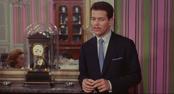 Movie still from “The Umbrellas of Cherbourg” (1964), directed by Jacques Demy – A man wearing a suit and tie sitting in front of a clock; Medium shot, Low angle