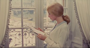 Movie still from “The Umbrellas of Cherbourg” (1964), directed by Jacques Demy – A woman standing in front of a window looking at a piece of paper; Medium shot, Low angle