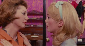 Movie still from “The Umbrellas of Cherbourg” (1964), directed by Jacques Demy – Two women talking to each other in front of a purple wall; Close Up shot, Over the shoulder angle