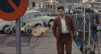 Movie still from “The Umbrellas of Cherbourg” (1964), directed by Jacques Demy – A man in a brown suit standing on a sidewalk; Wide shot, Over the shoulder angle