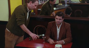 Movie still from “The Umbrellas of Cherbourg” (1964), directed by Jacques Demy – A man sitting at a table in front of a glass of wine; Medium shot, High angle