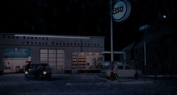 Movie still from “The Umbrellas of Cherbourg” (1964), directed by Jacques Demy – A gas station with cars parked in front of it at night; Extreme Wide shot, High angle