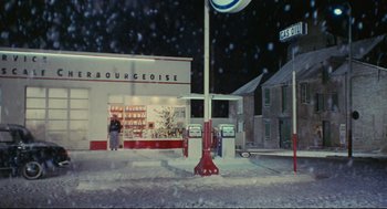 Movie still from “The Umbrellas of Cherbourg” (1964), directed by Jacques Demy – A gas station and gas pumps on a snowy day; Extreme Wide shot, High angle