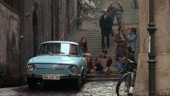 Movie still from “The Unbearable Lightness of Being” (1988), directed by Philip Kaufman – A group of people sitting on steps next to an old car; Wide shot, High angle