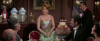 Movie still from “The Unsinkable Molly Brown” (1964), directed by Charles Walters – A woman standing in front of a cake on a table; Medium shot, Over the shoulder angle