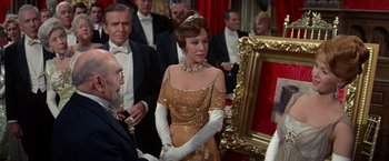 Movie still from “The Unsinkable Molly Brown” (1964), directed by Charles Walters – A woman in a gold dress standing in front of a group of people; Medium shot, Over the shoulder angle
