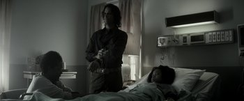 Movie still from “The Veil” (2016), directed by Phil Joanou – A man standing next to a woman laying in a hospital bed; Medium shot, Over the shoulder angle
