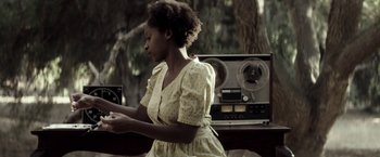 Movie still from “The Veil” (2016), directed by Phil Joanou – A woman in a yellow and white dress is standing in front of an old radio; Medium shot, Over the shoulder angle