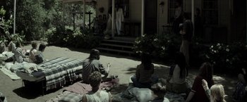 Movie still from “The Veil” (2016), directed by Phil Joanou – A group of people sitting on the ground in front of a house; Wide shot, High angle