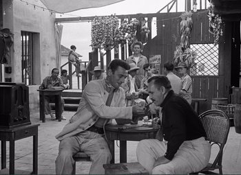 Movie still from “The Wages of Fear” (1953), directed by Henri-Georges Clouzot – Two men sitting at an outdoor table and one is holding his hand out to another man who is sitting at a table; Wide shot, High angle