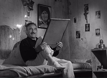 Movie still from “The Wages of Fear” (1953), directed by Henri-Georges Clouzot – An older man sitting on a bed reading a book; Medium shot, Low angle