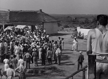 Movie still from “The Wages of Fear” (1953), directed by Henri-Georges Clouzot – A group of people standing in front of a building; Extreme Wide shot, Over the shoulder angle
