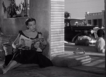 Movie still from “The Wages of Fear” (1953), directed by Henri-Georges Clouzot – A black and white photo of a woman sitting on the ground reading a magazine; Medium shot, High angle