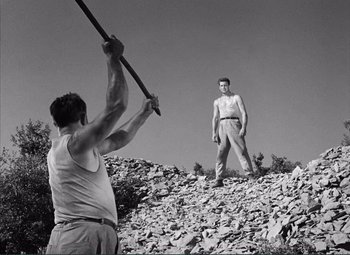Movie still from “The Wages of Fear” (1953), directed by Henri-Georges Clouzot – A man holding a baseball bat while standing on top of a pile of rocks; Wide shot, Low angle