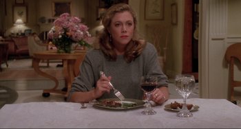 Movie still from “The War of the Roses” (1989), directed by Danny DeVito – A woman sitting at a table with a plate of food and a glass of wine in front of her; Medium shot, High angle