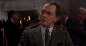 Movie still from “The War of the Roses” (1989), directed by Danny DeVito – An older man wearing a suit and tie in front of a fireplace; Close Up shot, Over the shoulder angle