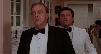Movie still from “The War of the Roses” (1989), directed by Danny DeVito – A man in a tuxedo standing next to another man in a suit; Medium shot, Low angle