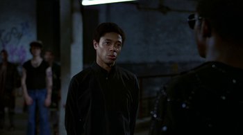 Movie still from “The Warriors” (1979), directed by Walter Hill – A man in a black shirt is standing in a dark room; Medium shot, Over the shoulder angle