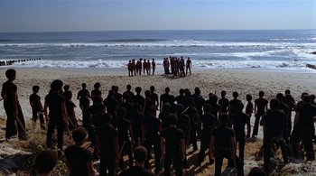 Movie still from “The Warriors” (1979), directed by Walter Hill – A group of people standing on top of a sandy beach next to the ocean; Extreme Wide shot, High angle