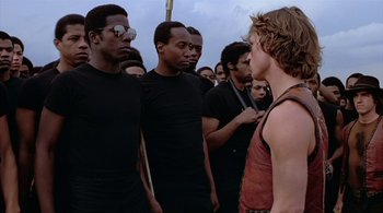 Movie still from “The Warriors” (1979), directed by Walter Hill – A group of men standing next to each other in front of a sky background; Medium shot, Over the shoulder angle