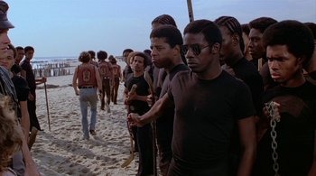 Movie still from “The Warriors” (1979), directed by Walter Hill – A group of men standing on top of a sandy beach; Medium shot, High angle