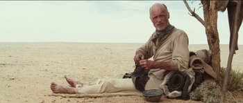 Movie still from “The Way Back” (2010), directed by Peter Weir – An older man sitting on the ground with a bowl of food; Medium shot, Low angle