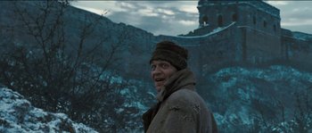 Movie still from “The Way Back” (2010), directed by Peter Weir – A man wearing a coat and hat standing in front of a wall; Medium shot, Low angle
