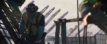 Movie still from “The Way Back” (2020), directed by Gavin O'Connor – A construction worker is wearing a hard hat and ear muffs while standing in front of a crane; Wide shot, Low angle