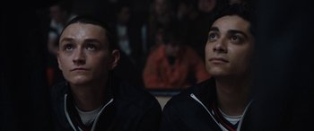 Movie still from “The Way Back” (2020), directed by Gavin O'Connor – Two young men sitting next to each other in front of a group of people; Close Up shot, Low angle