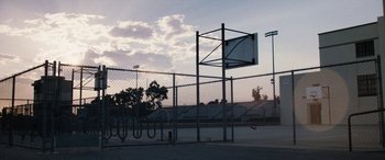 Movie still from “The Way Back” (2020), directed by Gavin O'Connor – A basketball hoop in a fenced in area; Extreme Wide shot, Low angle