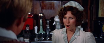 Movie still from “The Way We Were” (1973), directed by Sydney Pollack – A woman in a white dress and checkered hat in front of a stove; Close Up shot, Over the shoulder angle