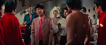 Movie still from “The Way of the Dragon” (1972), directed by Bruce Lee – A group of men standing around each other in a room; Medium shot, Over the shoulder angle