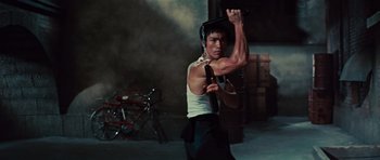Movie still from “The Way of the Dragon” (1972), directed by Bruce Lee – A man in a white tank top and black pants holding a gun; Medium shot, Low angle
