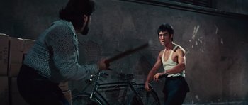 Movie still from “The Way of the Dragon” (1972), directed by Bruce Lee – A man holding a baseball bat in front of another man holding a bicycle; Medium shot, Over the shoulder angle