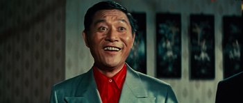 Movie still from “The Way of the Dragon” (1972), directed by Bruce Lee – An older man in a suit and a red shirt smiling; Close Up shot, Low angle