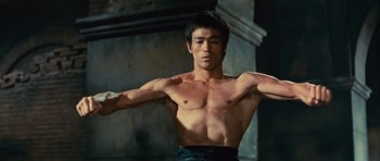 Movie still from “The Way of the Dragon” (1972), directed by Bruce Lee – A man with no shirt and no shirt is holding his arms out; Medium shot, Low angle