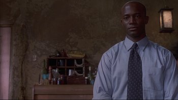 Movie still from “The Way of the Gun” (2000), directed by Christopher McQuarrie – A man standing in front of a wall with a shelf behind him; Medium shot, Low angle